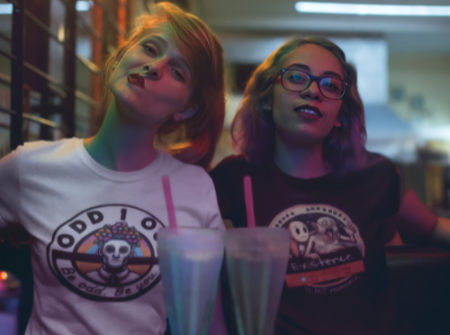 Two women sit in a cozy café wearing Odd 1 Out graphic t-shirts. The white shirt features the official Odd 1 Out logo with skull design and “Be odd, be you” tagline. A bold visual of authentic streetwear and unapologetic style.